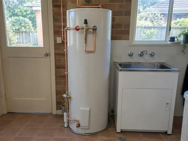 Hot water cylinder with copper pipework in a laundry beside a sink and brick wall in Hornsby